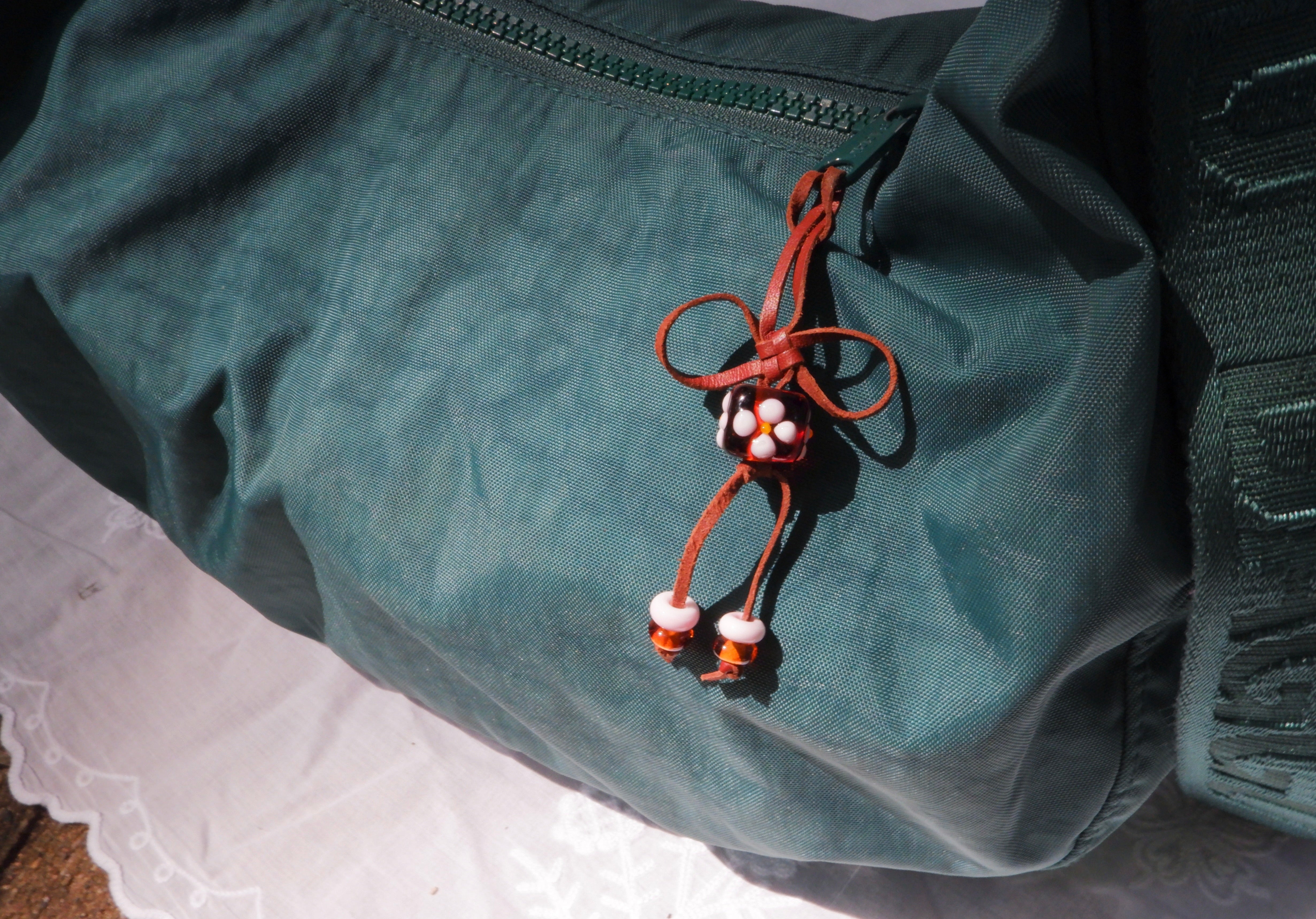 A dark teal-green nylon bag with a brown cord tassel charm featuring white and topaz floral beads, resting on a white lace-trimmed surface.