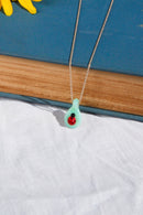 A delicate silver chain necklace with a small mint green glass pendant featuring a red ladybug, displayed on a blue book over a white fabric background with yellow flowers.