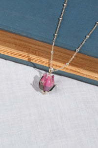 A delicate silver beaded chain necklace featuring a small pink glass flower bud pendant, styled on a blue book resting on a white textured fabric surface.